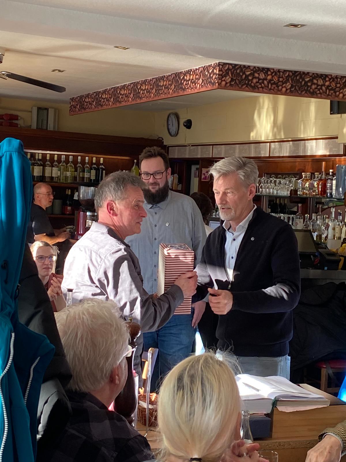 Christoph überreicht Udo ein Geschenk. Da hinter steht Nils. Im Hintergrund ist ein Lokal zu erkennen.