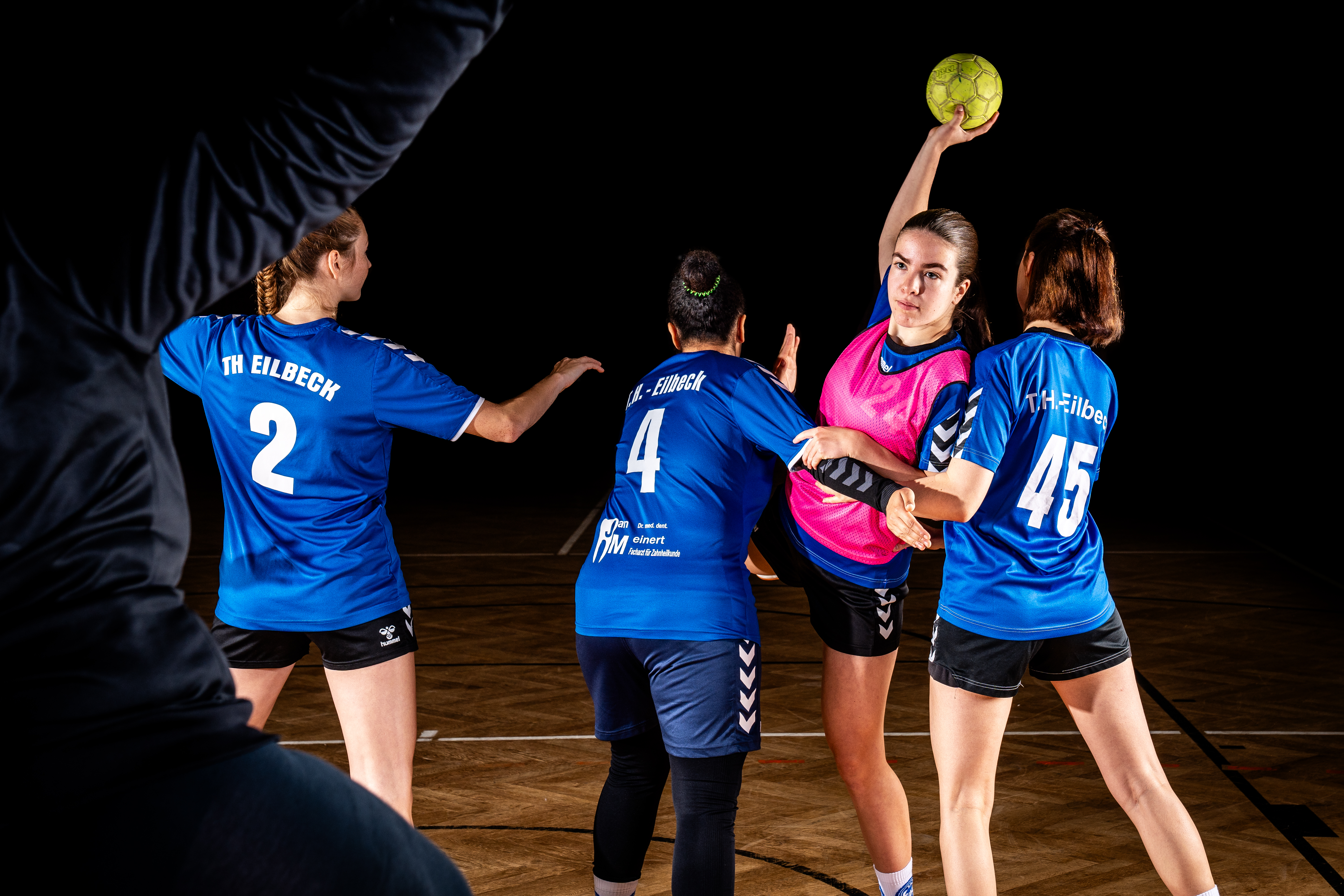 Das Bild zeigt vier weibliche Handballspielerinnen in blauen Trikots mit der Aufschrift "TH Eilbeck" und Nummern auf dem Rücken. Eine Spielerin in einem pinken Trikot wirft den Ball, während die anderen Spielerinnen in Verteidigungsstellungen um sie herum stehen. Ein Teil eines Trainers oder Betreuers ist im Vordergrund zu sehen. Der Hintergrund ist dunkel, der Fokus liegt auf den Spielerinnen. Die Szene scheint ein Spiel oder Training im Handball zu zeigen.