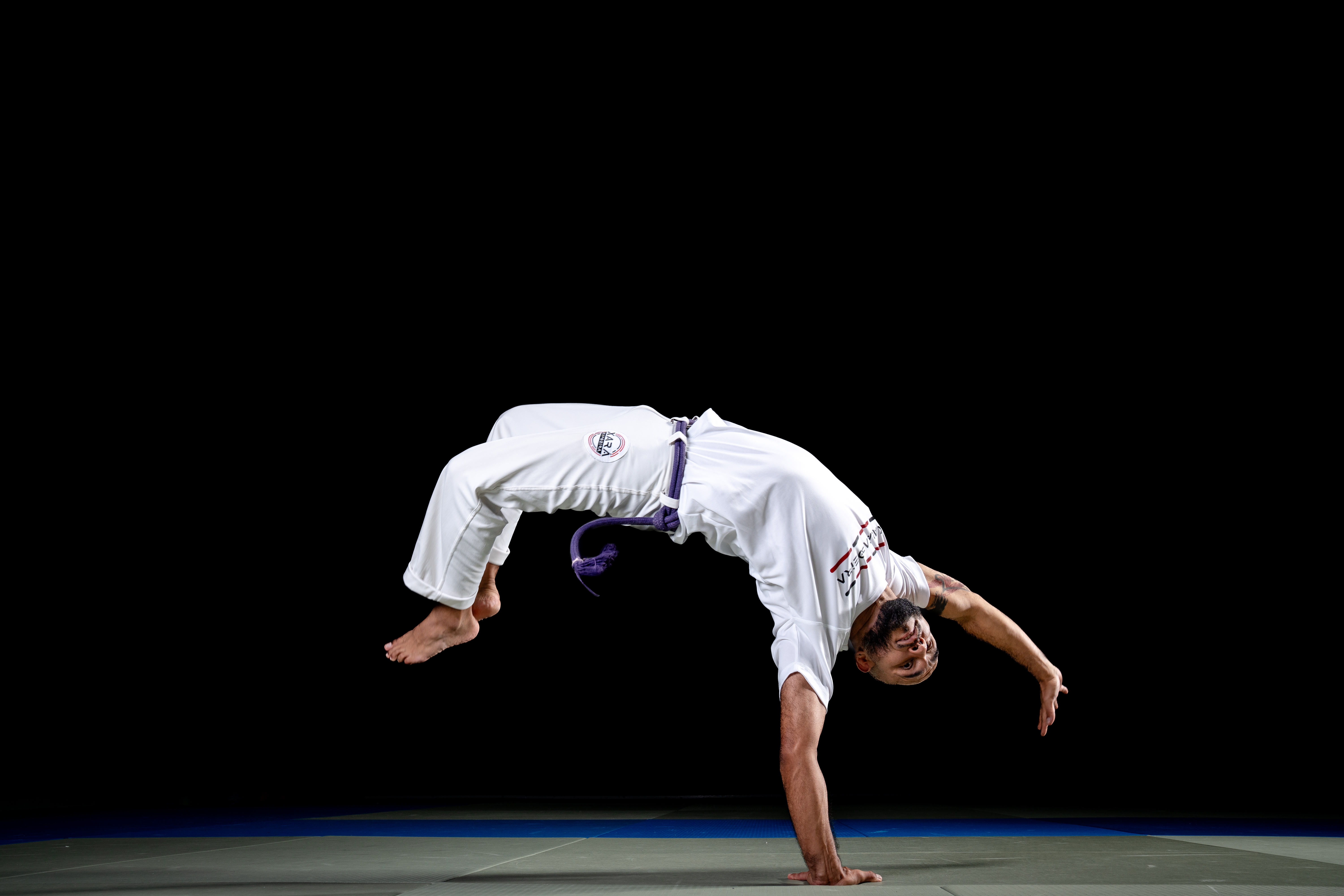 Ein Sportler (Capoeira) in weißem Shirt und weißer Hose mit schwarzer Kordel steht auf eine Hand. Der Hintergrund ist dunkel. Unten sind Budomatten erkennbar.