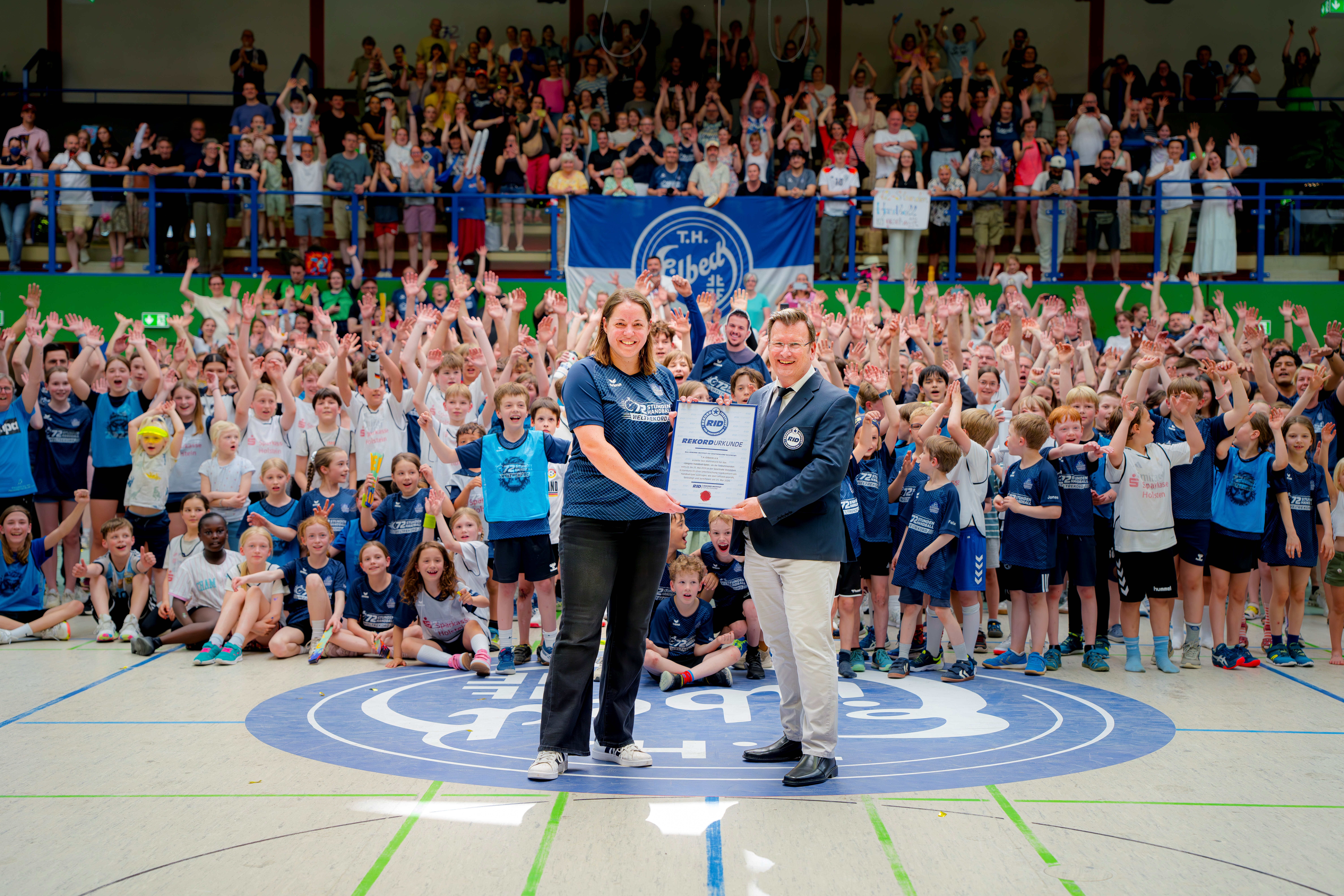 Große Gruppe von Kindern und Erwachsenen in einer Sporthalle, viele mit erhobenen Armen. Im Vordergrund stehen zwei Personen auf dem Hallenboden mit Vereinslogo und halten gemeinsam eine gerahmte Urkunde. Hinter ihnen stehen zahlreiche Kinder in Sportkleidung sowie Zuschauer auf Tribünen; im Hintergrund ist ein großes Banner mit der Aufschrift „T.H. Eilbeck“ sichtbar.