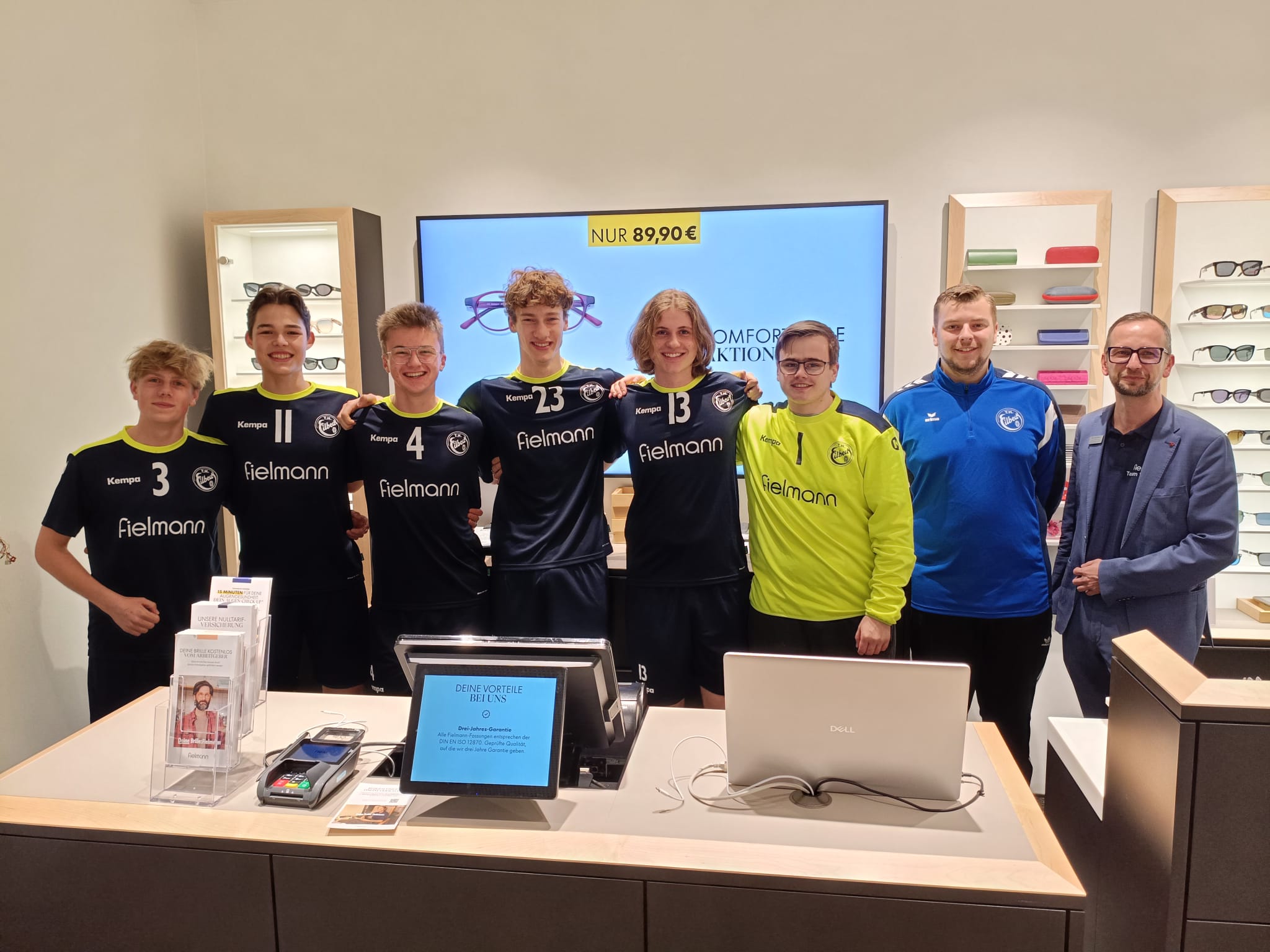 Fünf Spieler in schwarzen Trikots mit gelbem Rand und der Aufschrift Fielmann stehen neben ihrem Torwart in einem gelben Trikot mit schwarzem Rand. Mit auf dem Bild sind auch ihr Trainer in blauer Trainingsjacke sowie ein Mann im Anzug (Niederlassungsleiter der Fielmann-Filiale).