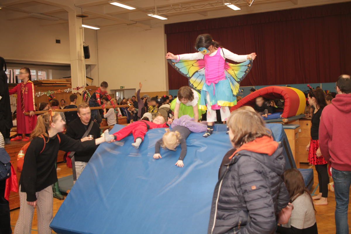 Kinder turnen im Kstüm über einen großen Mattenberg. Im Hintergrund und neben dem Mattenberg sind viele Kinder und Erwachsene zu erkennen.